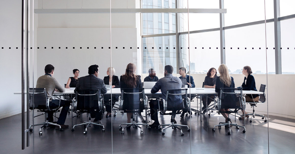 A crowded and energetic conference room in a futuristic building. Roughly a dozen interested employees engage in discussion. 