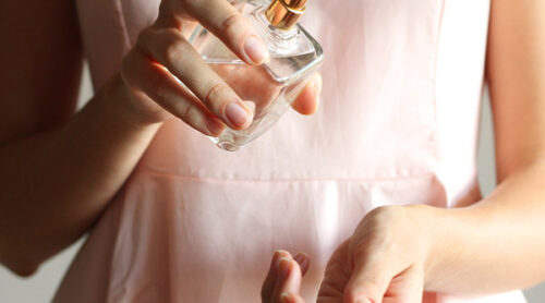 A woman in a pink shirt applies perfume on her wrist.