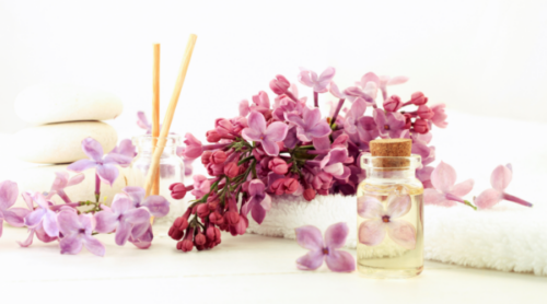 Fragrance bottles and purple floral arrangements on a white table and background.